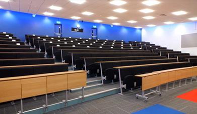 Empty lecture room with rows of seats and desks facing a screen. 