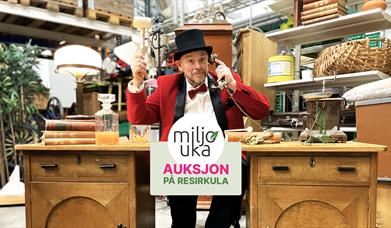 Mann i flosshatt holder hammer, mens han sitter i telefonen bak et antikk skrivebord. Miljøuka-logo og tittel "Auksjon på Resirkula" i grønne og rosa farger.