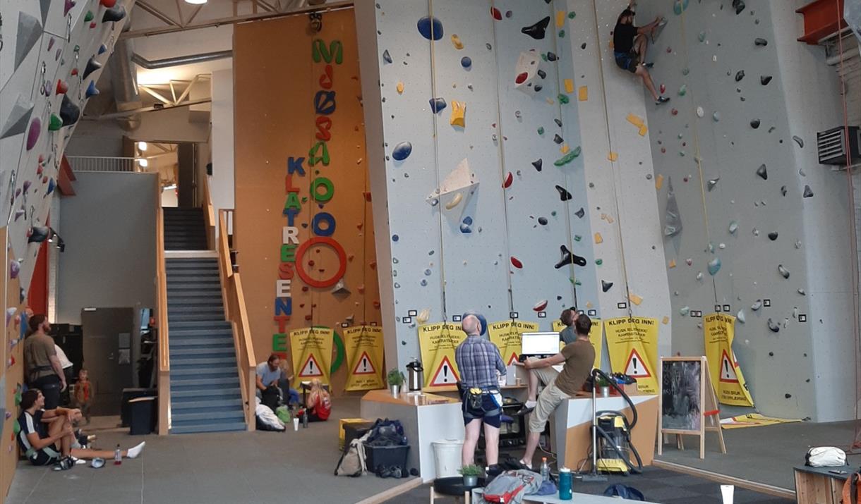 An indoor climbing facility with tall, light-colored climbing walls, multiple auto-belay lines, and routes marked by colorful holds. Climbing gear and mats are on the floor, and several people are visible in the background moving around the space.