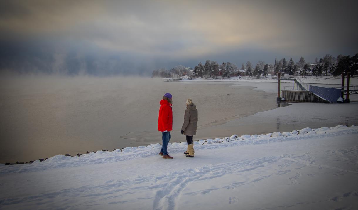 Two people stand at the edge of a snow-covered lakeside with soft winter sunlight and light clouds overhead.