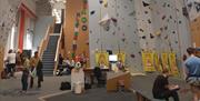A section of an indoor climbing center with tall walls covered in colorful climbing holds. There are open floor areas and a few people visible in the