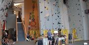An indoor climbing facility with tall, light-colored climbing walls, multiple auto-belay lines, and routes marked by colorful holds. Climbing gear and mats are on the floor, and several people are visible in the background moving around the space.