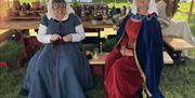 Two people dressed in historical medieval clothing are seated at a wooden table in an outdoor medieval‑themed setting. The table is filled with potter