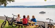 Four people sit at a picnic table by the water, with bicycles parked nearby.