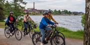 A group of people ride bicycles along a path by the water with the town visible in the background.