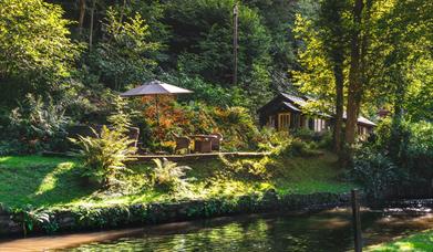 Picture of cottage and outdoor seating from across the river. Sunlight seen between trees.