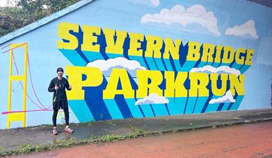 parkrun runner standing next to Severn Bridge Parkrun sign - bright and massive painted on wall at start of parkrun in chepstow
