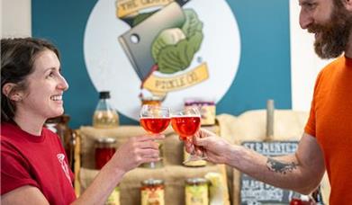 Brunette man and woman on either side of picture, in front of a display of fermented foods cheers-ing with a glass of pink kombucha