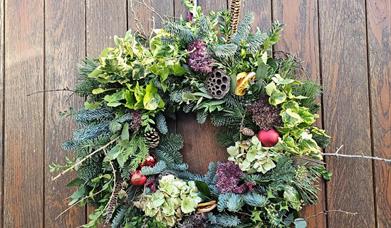 A natural Christmas wreath featuring evergreens, hydrangea blooms, pinecones, feathers, and red ornaments, hanging on a rustic wooden door with a fest