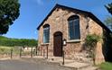 View of chapel in the sunshine from outside looking at the door, car park, steps and a ramp with a handrail are in front. The chapel is surrounded by