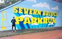 parkrun runner standing next to Severn Bridge Parkrun sign - bright and massive painted on wall at start of parkrun in chepstow