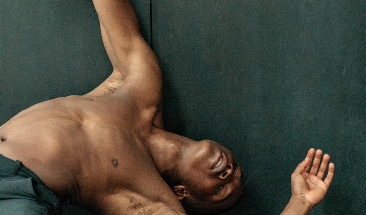 A shirtless man wearing dark pants leans back against a dark wall with his arms outstretched and head tilted, appearing to be in a dance or stretching