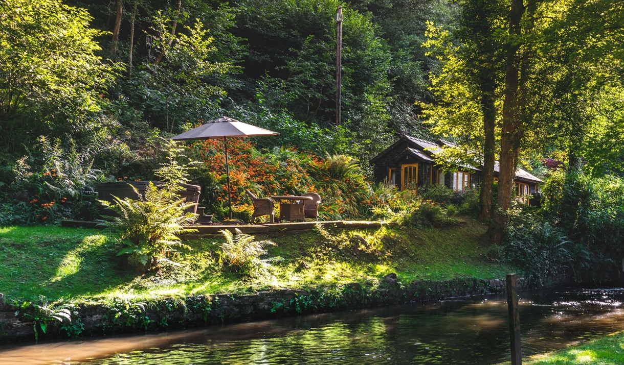 Picture of cottage and outdoor seating from across the river. Sunlight seen between trees.