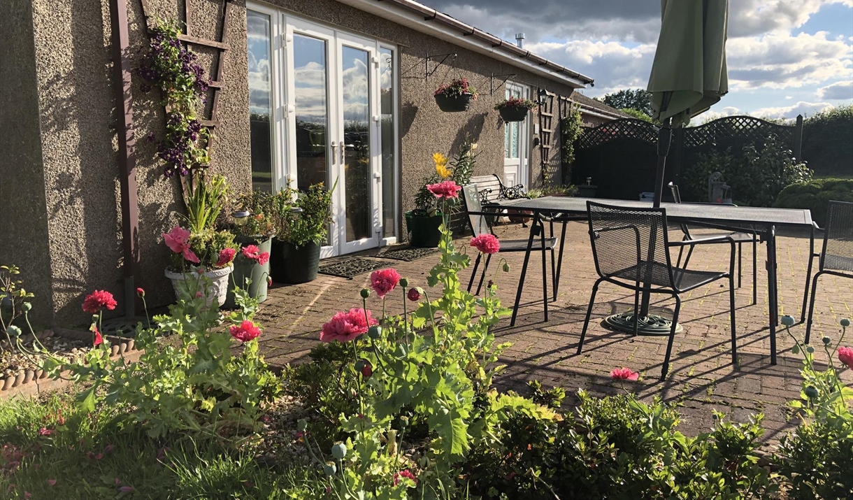 Front view of Blackthorn Lodge showing the lounge patio doors, front door and patio garden area with garden furniture including a table, 4 chairs, par