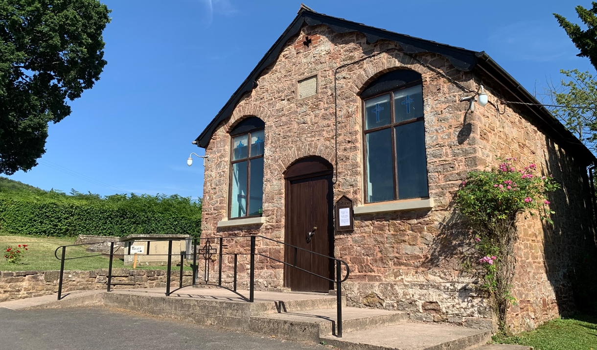 View of chapel in the sunshine from outside looking at the door, car park, steps and a ramp with a handrail are in front. The chapel is surrounded by