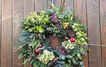 A natural Christmas wreath featuring evergreens, hydrangea blooms, pinecones, feathers, and red ornaments, hanging on a rustic wooden door with a fest