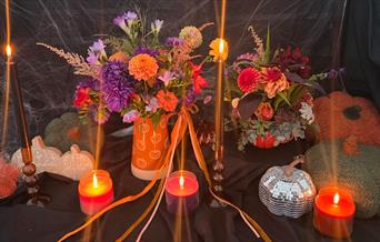 Halloween-themed floral display with vibrant autumn flowers arranged in pumpkin containers, surrounded by glowing candles, black taper holders, and so