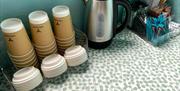 Table with green spotty tablecloth and facilities to make refreshments. A kettle, takeaway cups and lids are in the foreground, with tea, coffee, suga