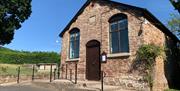 View of chapel in the sunshine from outside looking at the door, car park, steps and a ramp with a handrail are in front. The chapel is surrounded by