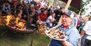 abergavenny food festival ladys smiles and makes silly face whilst holding a plate of food in the fire pit cooking area with crowds in abergavenny cas