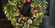 A festive Christmas wreath made of lush green foliage, red berries, cinnamon sticks, dried orange slices, and red baubles, hanging on a glossy black w