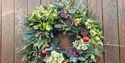A natural Christmas wreath featuring evergreens, hydrangea blooms, pinecones, feathers, and red ornaments, hanging on a rustic wooden door with a fest