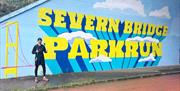 parkrun runner standing next to Severn Bridge Parkrun sign - bright and massive painted on wall at start of parkrun in chepstow
