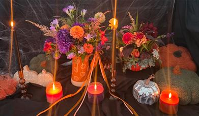 Halloween-themed floral display with vibrant autumn flowers arranged in pumpkin containers, surrounded by glowing candles, black taper holders, and so