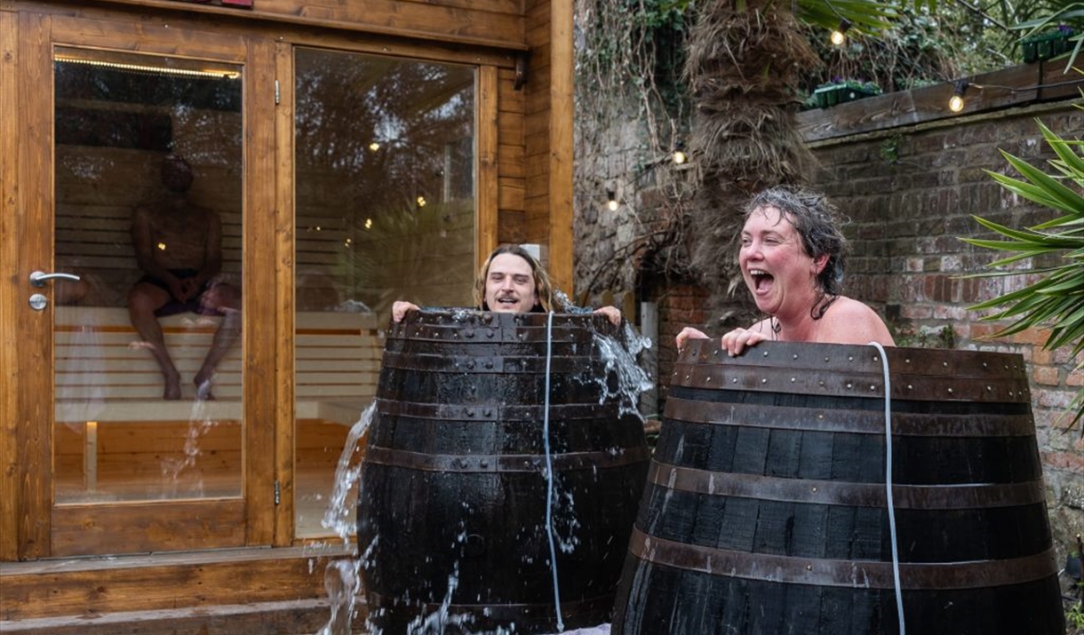 Woman and man laugh in cold plunge as cold water spills out of cold plunge barrel in Stone Rock Lodge Woman and man laugh in cold plunge as cold water spills out of cold plunge barrel in Stone Rock Lodge