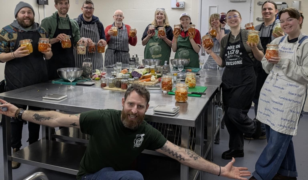 A group of people gathered around a table holding jars of fermented foods and the instructor at the front looking happy A group of people gathered around a table holding jars of fermented foods and the instructor at the front looking happy