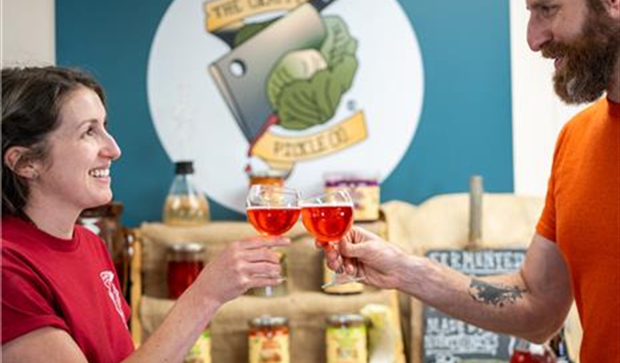 Brunette man and woman on either side of picture, in front of a display of fermented foods cheers-ing with a glass of pink kombucha Brunette man and woman on either side of picture, in front of a display of fermented foods cheers-ing with a glass of pink kombucha