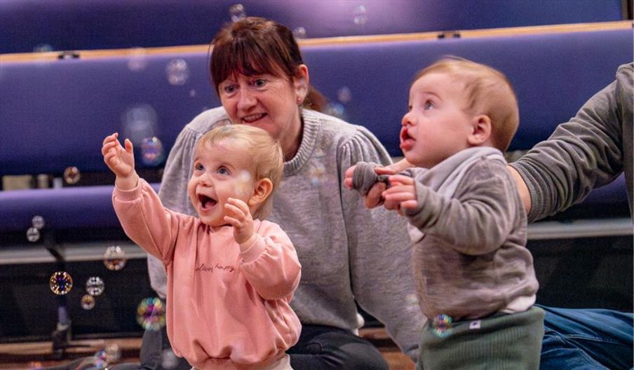 A baby in a pink jumper looks up in awe at a colourful toy held above their head by a woman wearing a patterned shirt and jeans. She kneels on the flo