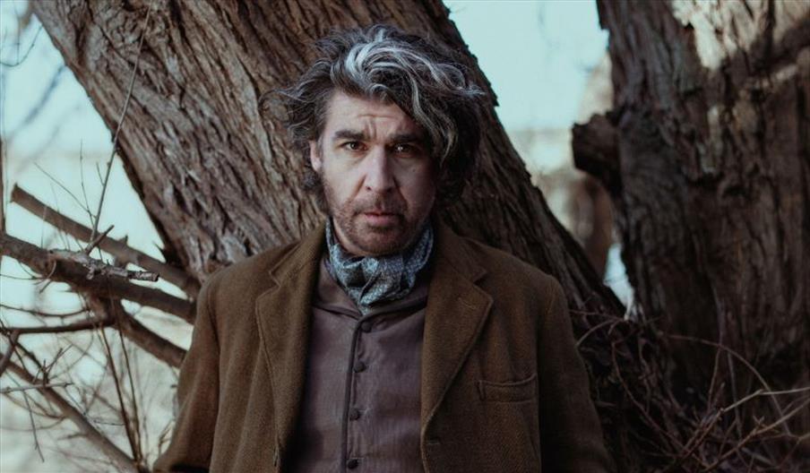 A man is stood in front of a wildly defined tree. He has wavy, black and grey hair with a deep side part. He is wearing a chocolate brown waistcoat, a