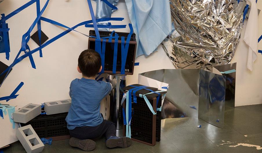 A child plays with blue tape and silver materials in a gallery.