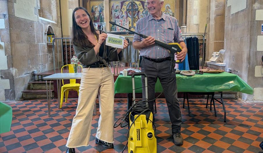 one person holding up a sign that reads "Fixed" and one person holding a Karcher pressure washer that has just been fixed at The Fixery.