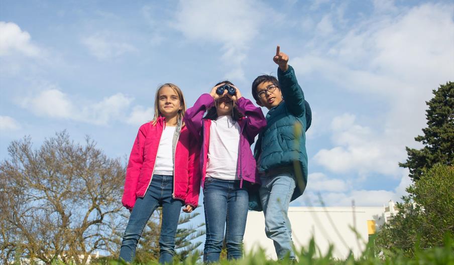 Three children, one of them pointing at something and the other looking at it with a pair of binoculars