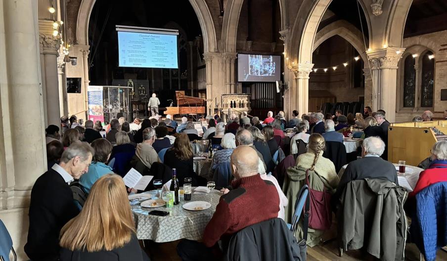 A large group of people are sitting around tables listening to a classical concert in a beautiful old church