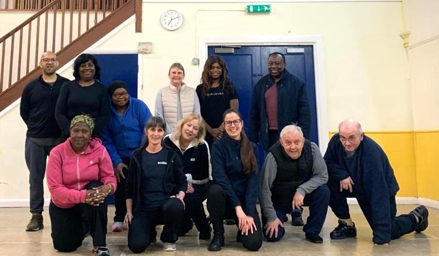 A diverse group of adults in a gym, smiling together, includes a personal trainer and a nutritionist.
