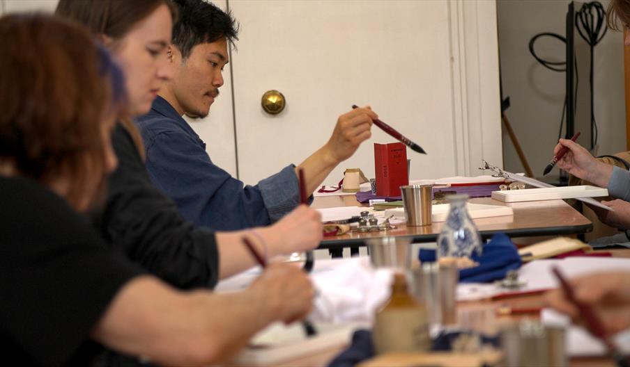 A group of artists , sitting at tables and holding mop-brushes, focus on their artworks at an Artanda workshop.