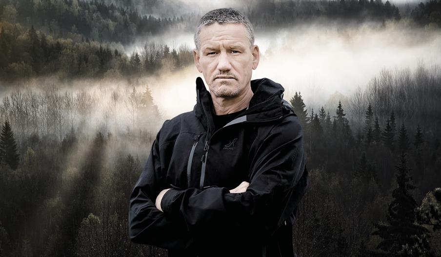 Mark Billingham stands facing the camera with his arms crossed and a serious expression on his face. He is against a backdrop of a misty mountain fore