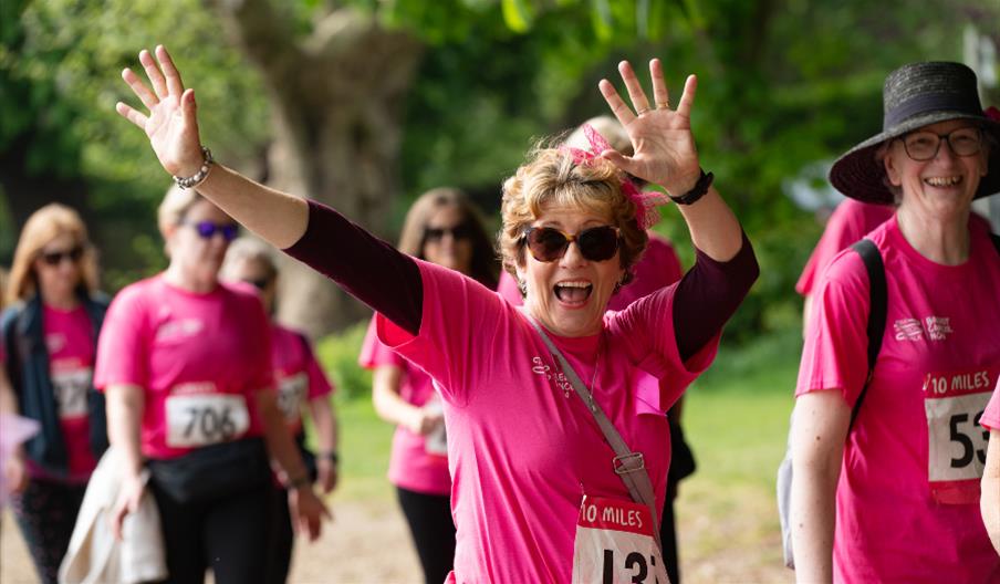 Woman wearing sunglasses and wearing a Breast Cancer Now T-shirt has her arms in the air and looks very happy. She is surrounded by other smiling wome