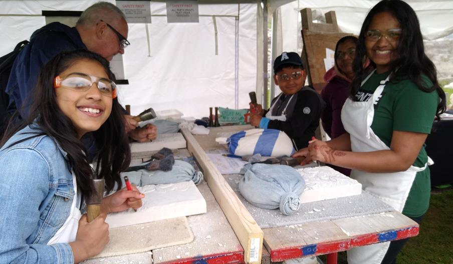 An image of five people, two children and three adults, carving into stone inside a tent wearing protective eyewear.