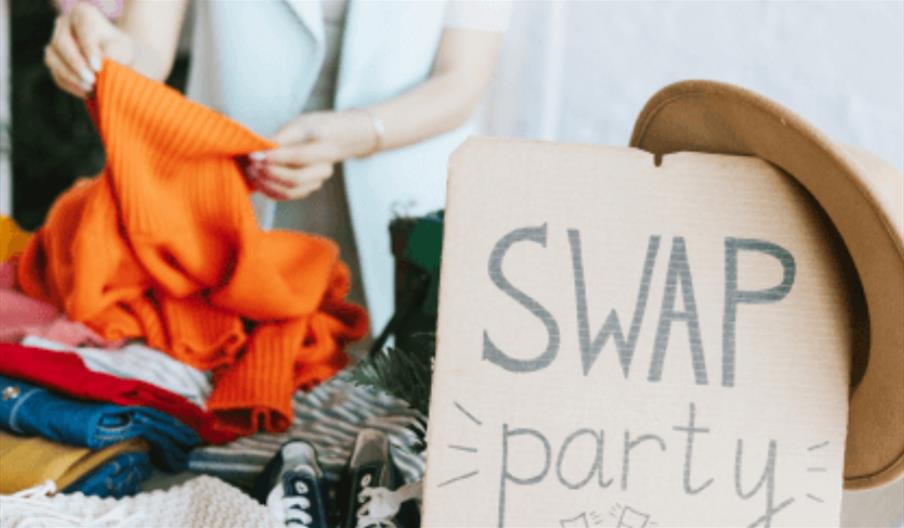 A poster with Swap Party written on cardboard in focus with a pile of folded clothes behind as a pair of hands browses through them.