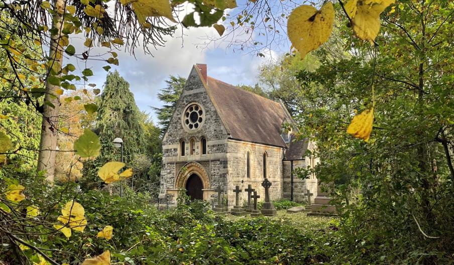 small gothic style chapel set in an old burial ground surrounded by nature. The image is taken on a sunny day through the amber leaves of a tree.
