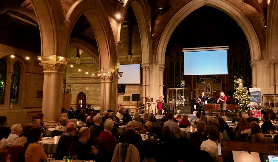 An audience sits at round tables in a festively lit church. At the front, musicians are playing a grand piano and a clarinet.