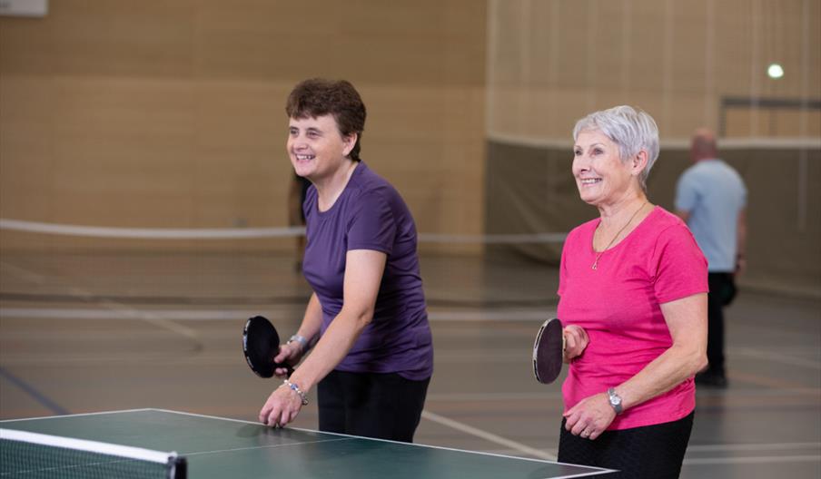 An image of two women with Parkinson's taking part in a friendly table tennis match together. One woman is wearing a purple top with black trousers an