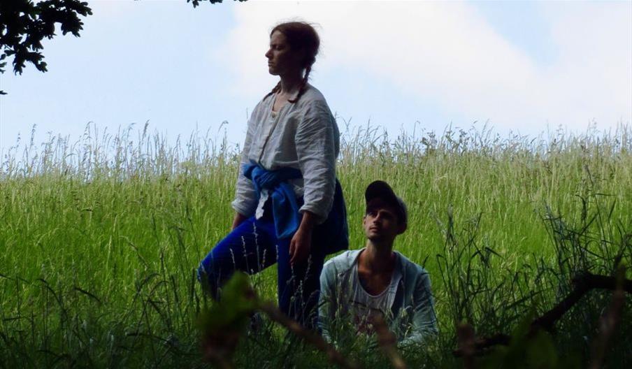 A standing woman and a seated man both with pale skin occupy the shady long grass beneath an oak tree. In the background is the brow of a grassy hill