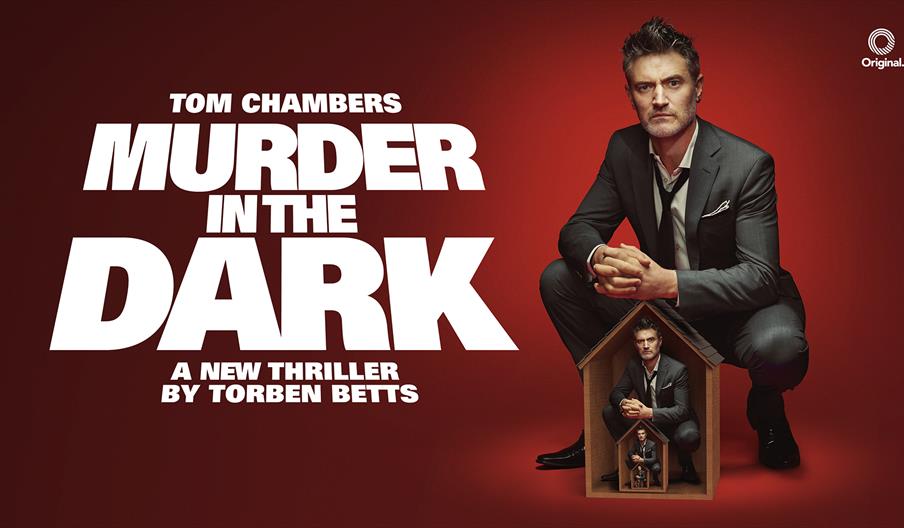 Poster for Murder in the Dark. On a dark red background, Tom Chambers is in a suit with the tie and collar loosened. He crouches over a small wooden h
