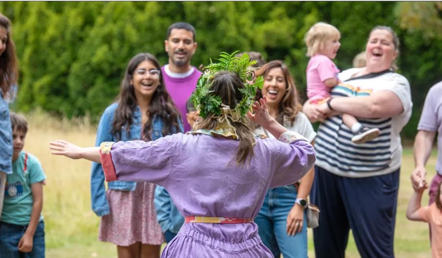 A woman in a purple jumpsuit wearing a crown made of leaves, stands with her back to the camera, gestures to laughing families