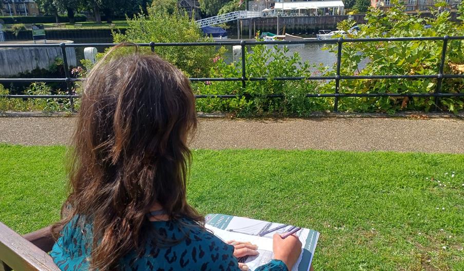 Image of a woman with long brown hair sat sketching near the Thames, taken from over her shoulder